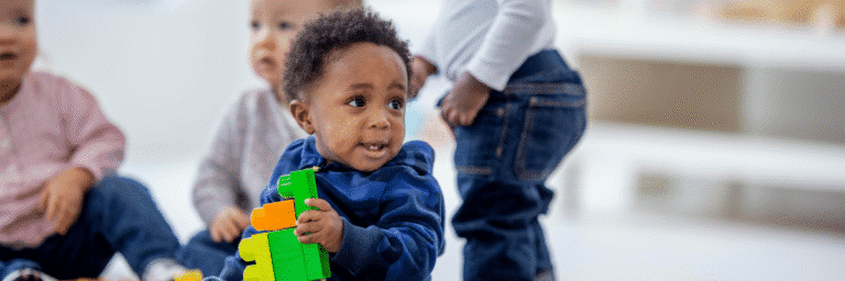 Toddler holding toy while adjusting to new preschool environment with other children in background